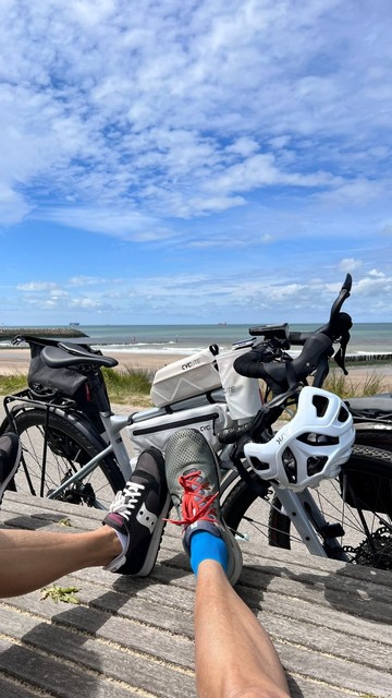 Zwei Füsse vor einem bepackten grauen Fahrrad, dahinter Strand und Meer.